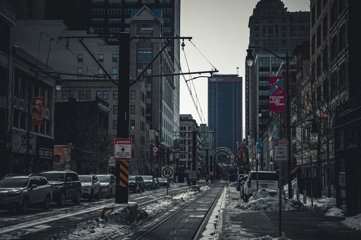 Snow-covered urban street with tram tracks, tall buildings, and overcast winter sky in city.