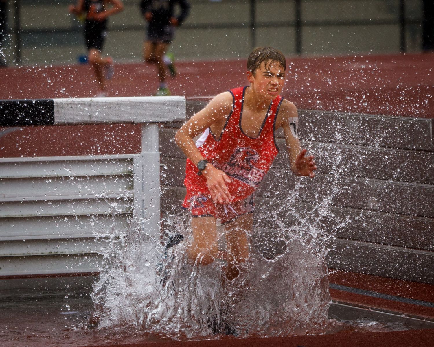 Male track athlete splashes through the water barrier during a steeplechase race on a red track.