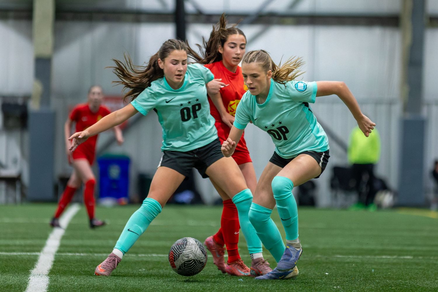 Three girls in teal and red uniforms compete for the ball during an indoor youth soccer match.