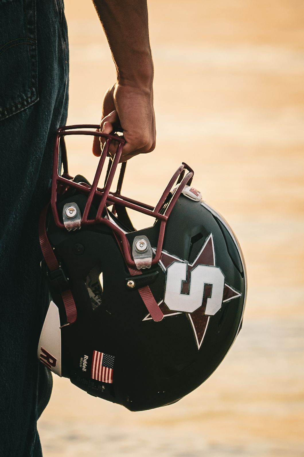 Hand holding a black and maroon football helmet with a star logo and American flag decal.