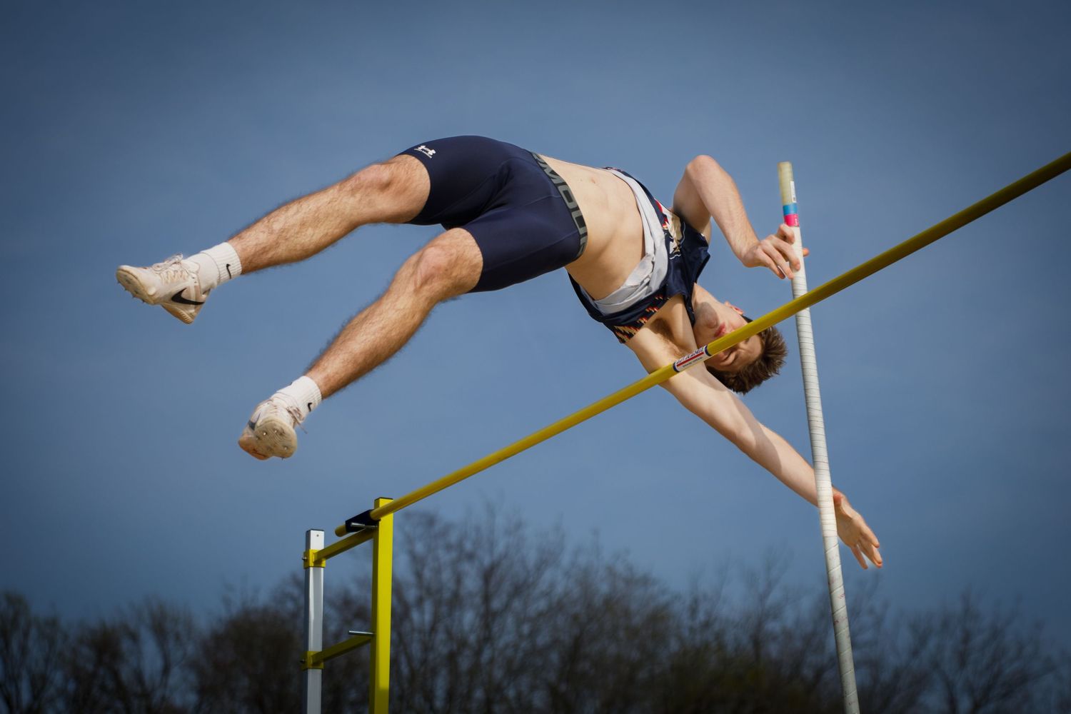 A male pole vaulter clears the bar in a dramatic arched position against a blue sky backdrop.
