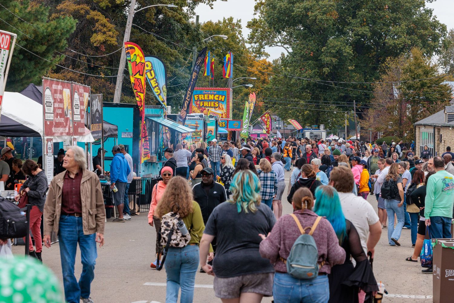 Large crowd of visitors strolling past colorful vendor booths and banners at a lively outdoor fair or festival on a sunny day.