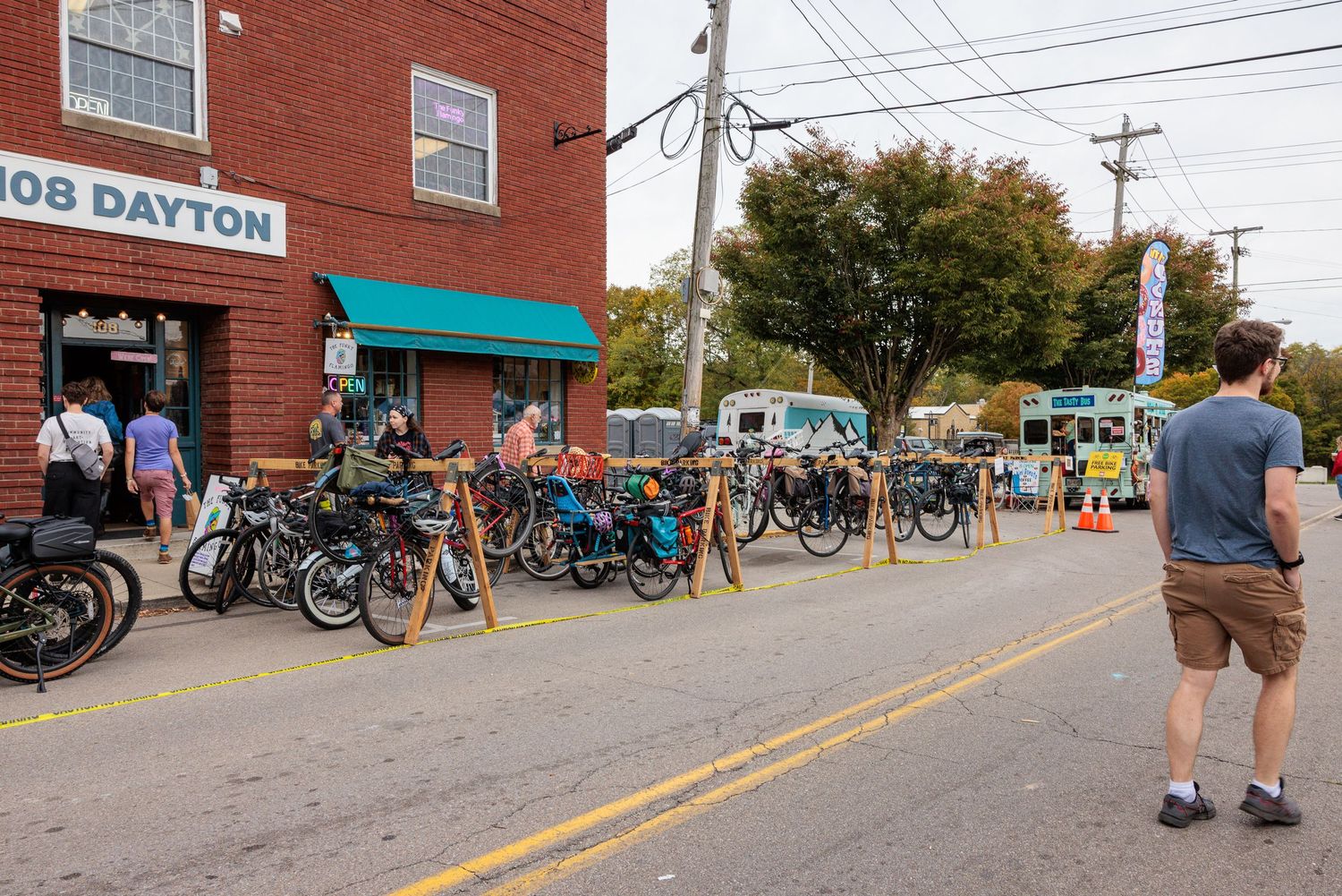 Man in shorts stands on a Dayton street facing a row of parked bicycles outside a brick building with a teal awning and food trucks nearby.