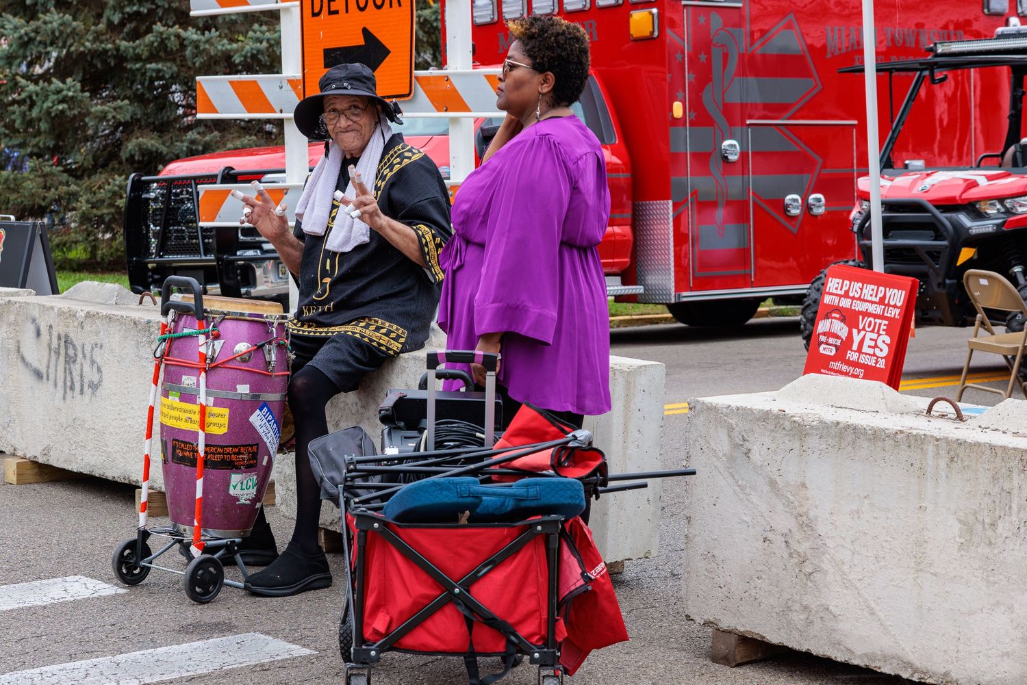 Two people in colorful clothing and hats stand beside rolling carts near concrete barriers and a red fire truck on a city street.