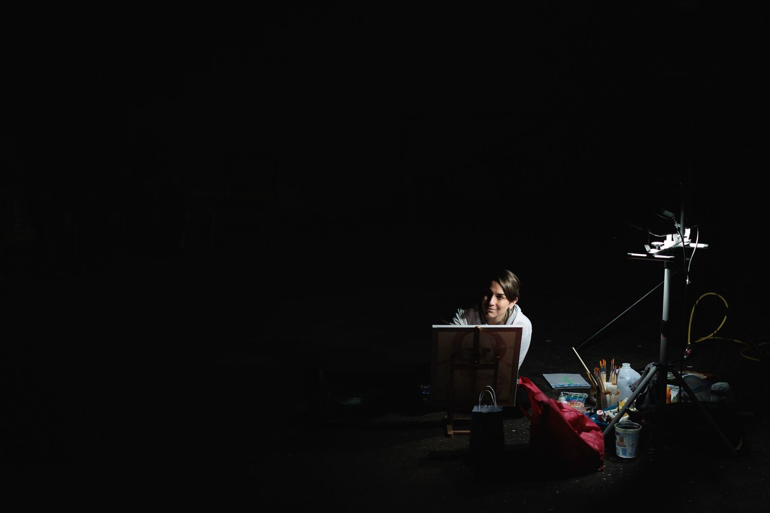 Low-light photo of a person seated at an easel, illuminated by a single light source