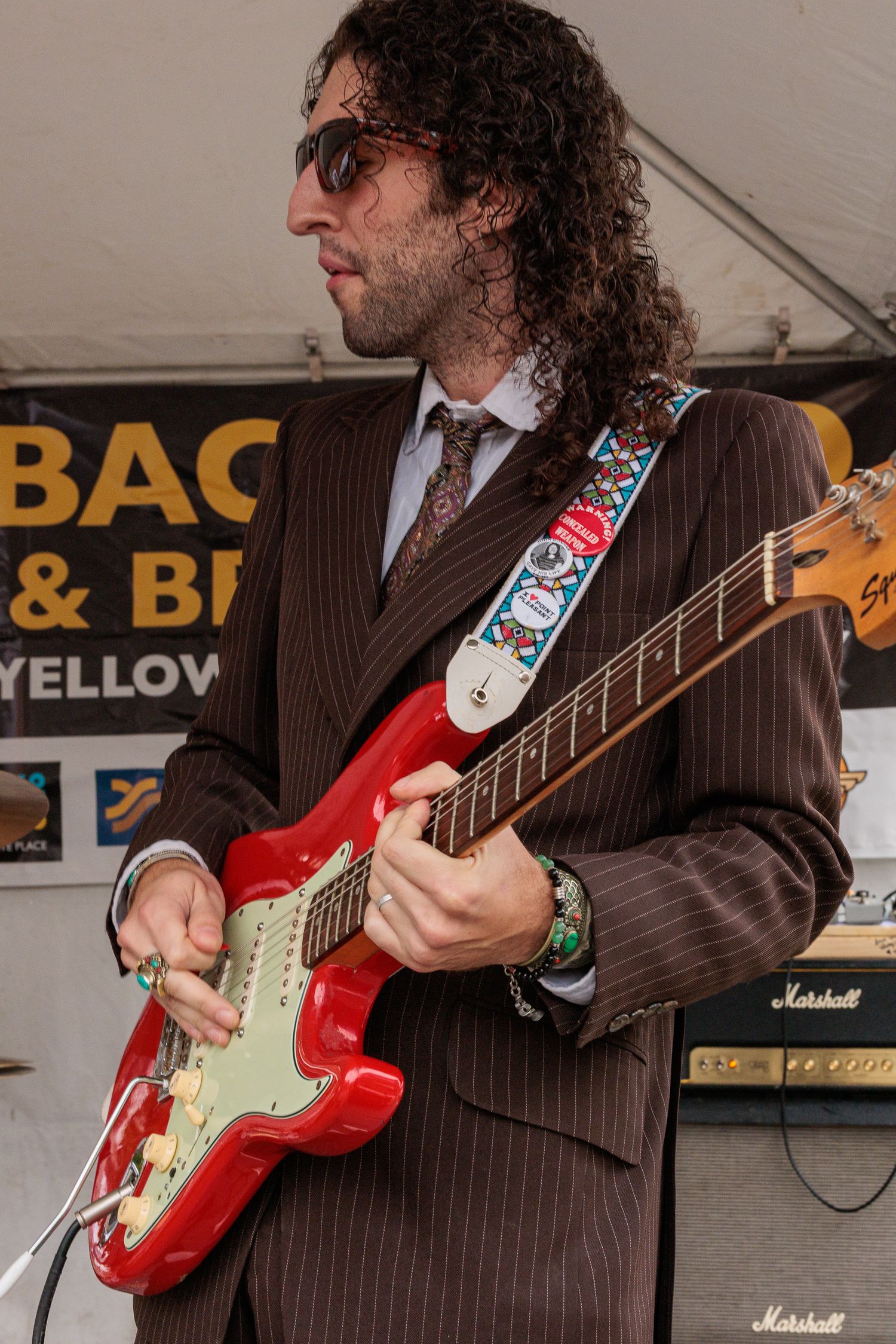 Guitarist with curly hair and sunglasses plays a red electric guitar on stage in a pinstripe suit, with Marshall amplifiers behind him.