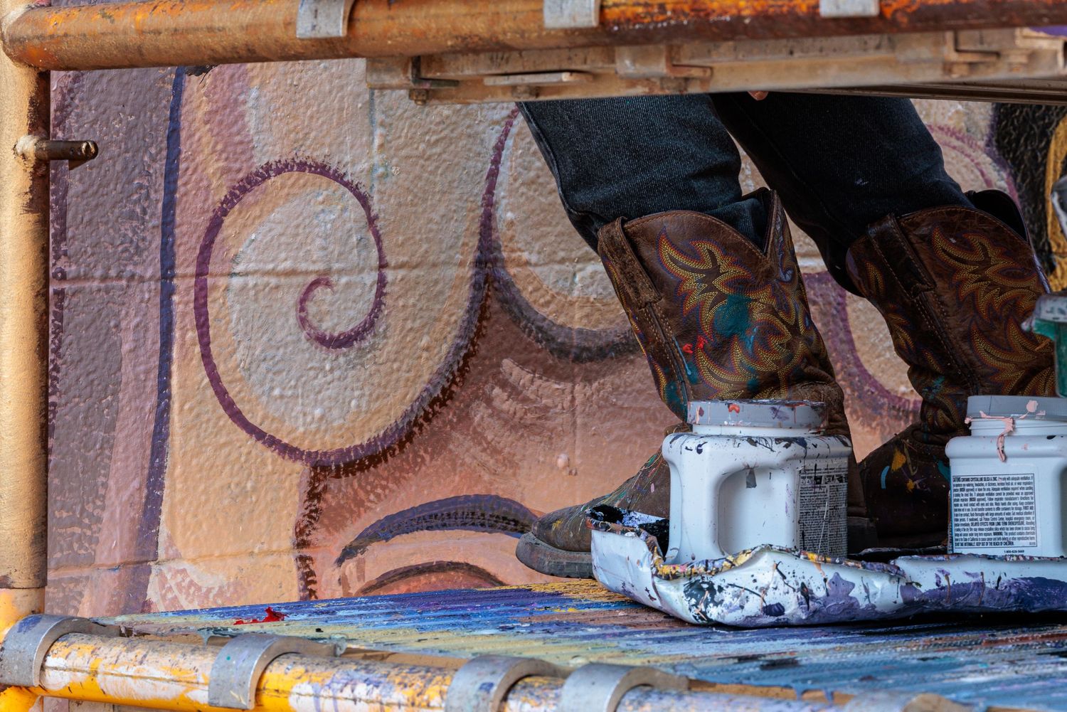 Close-up of paint buckets and boots at the base of scaffolding, spiral mural detail in background