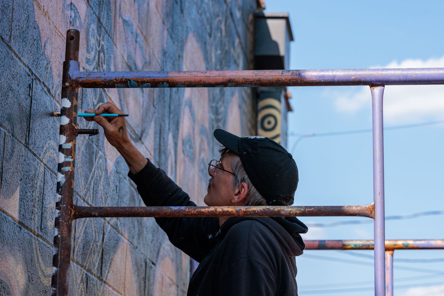 Muralist Pierre Nagley painting from scaffolding against a clear blue sky