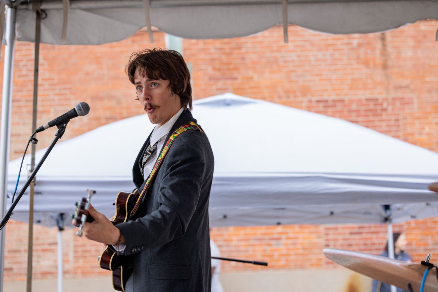 Musician in a dark suit plays an acoustic guitar and sings into a microphone beneath a white tent in front of a brick building.