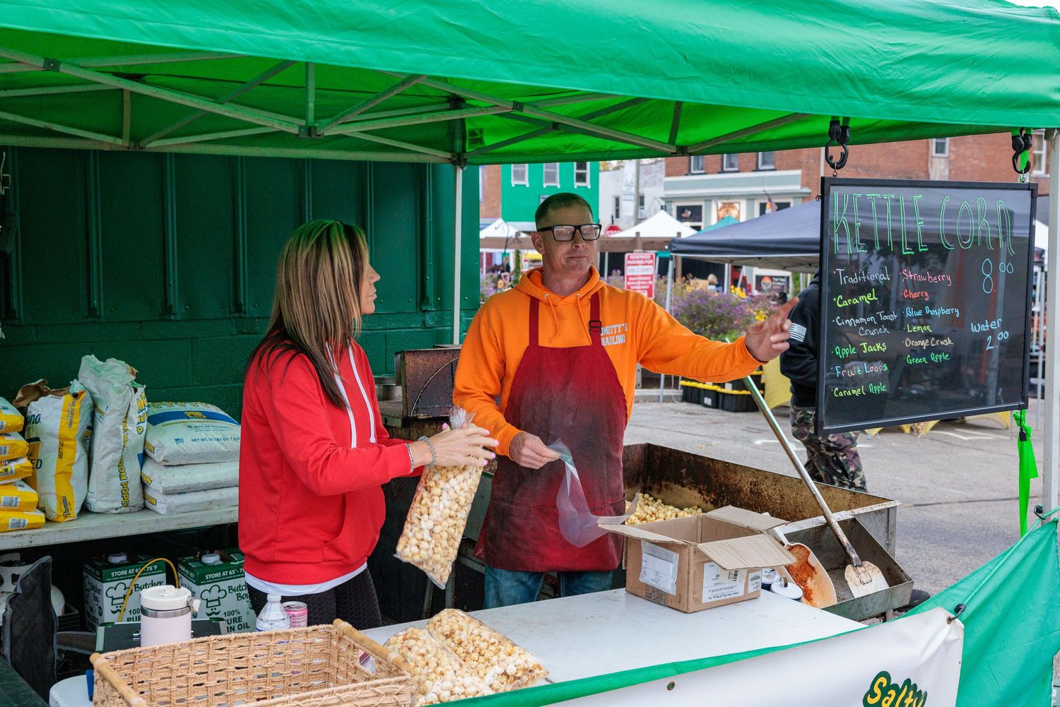 Smiling vendor in an orange hoodie and apron serves snacks to a woman in a red jacket at a green-canopied outdoor food market stall.
