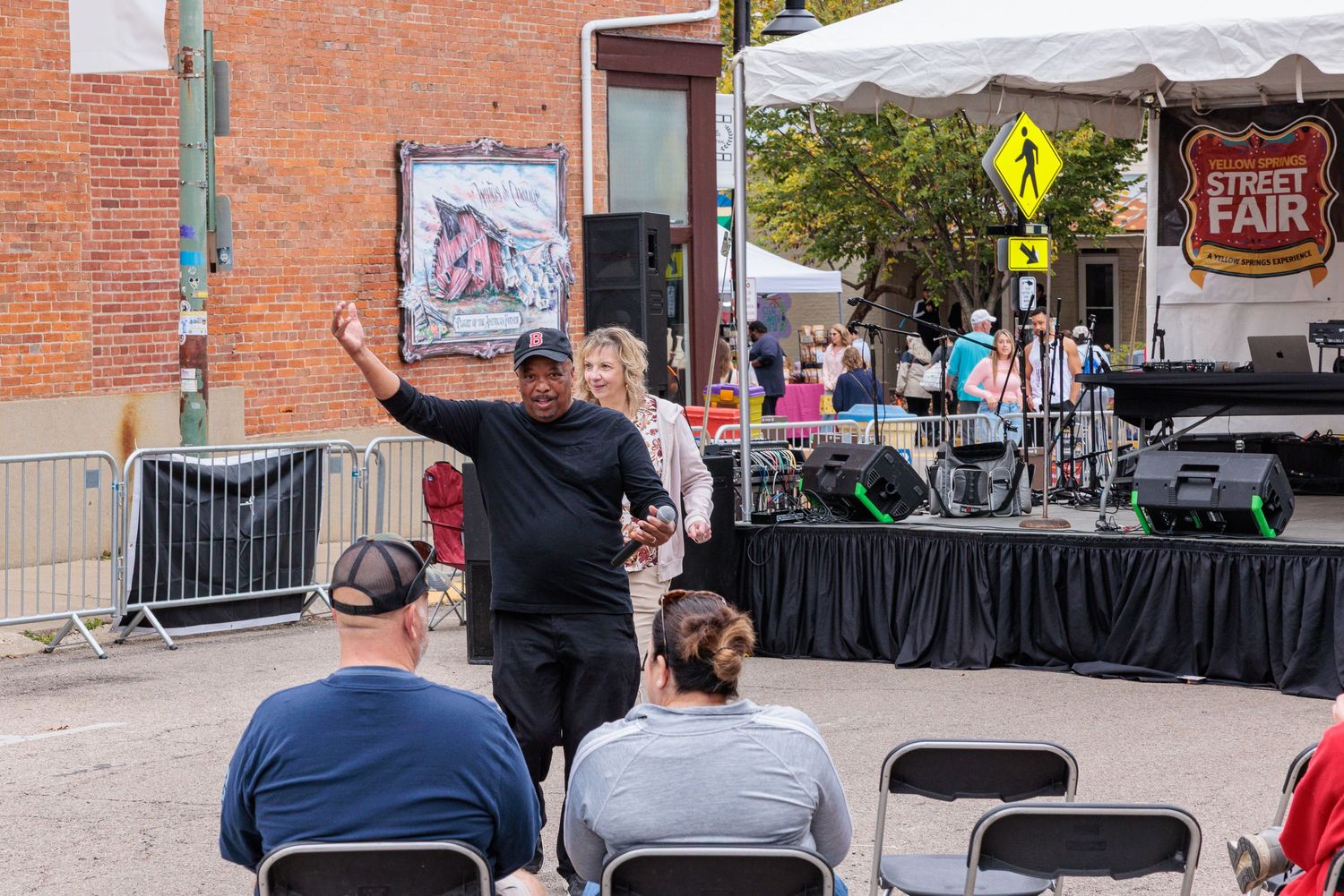 DJ in black cap and shirt gestures while speaking near an outdoor stage with speakers at a street fair as seated spectators watch.