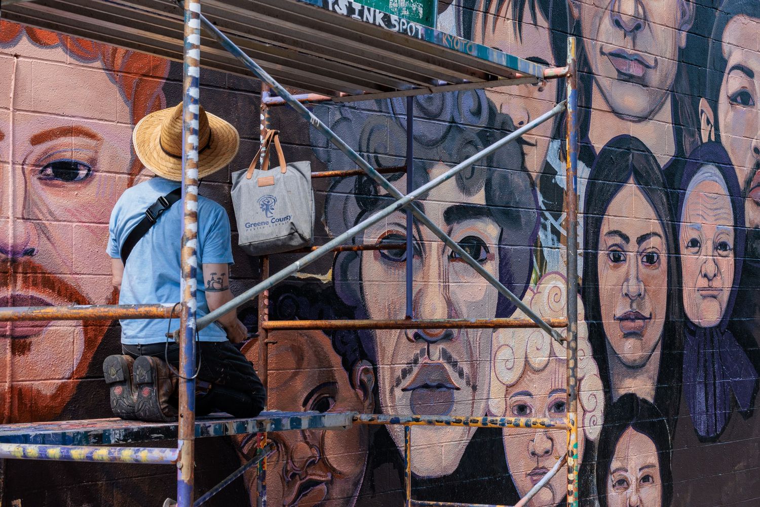 Muralist Pierre Nagley in a sun hat painting a row of portrait faces on scaffolding