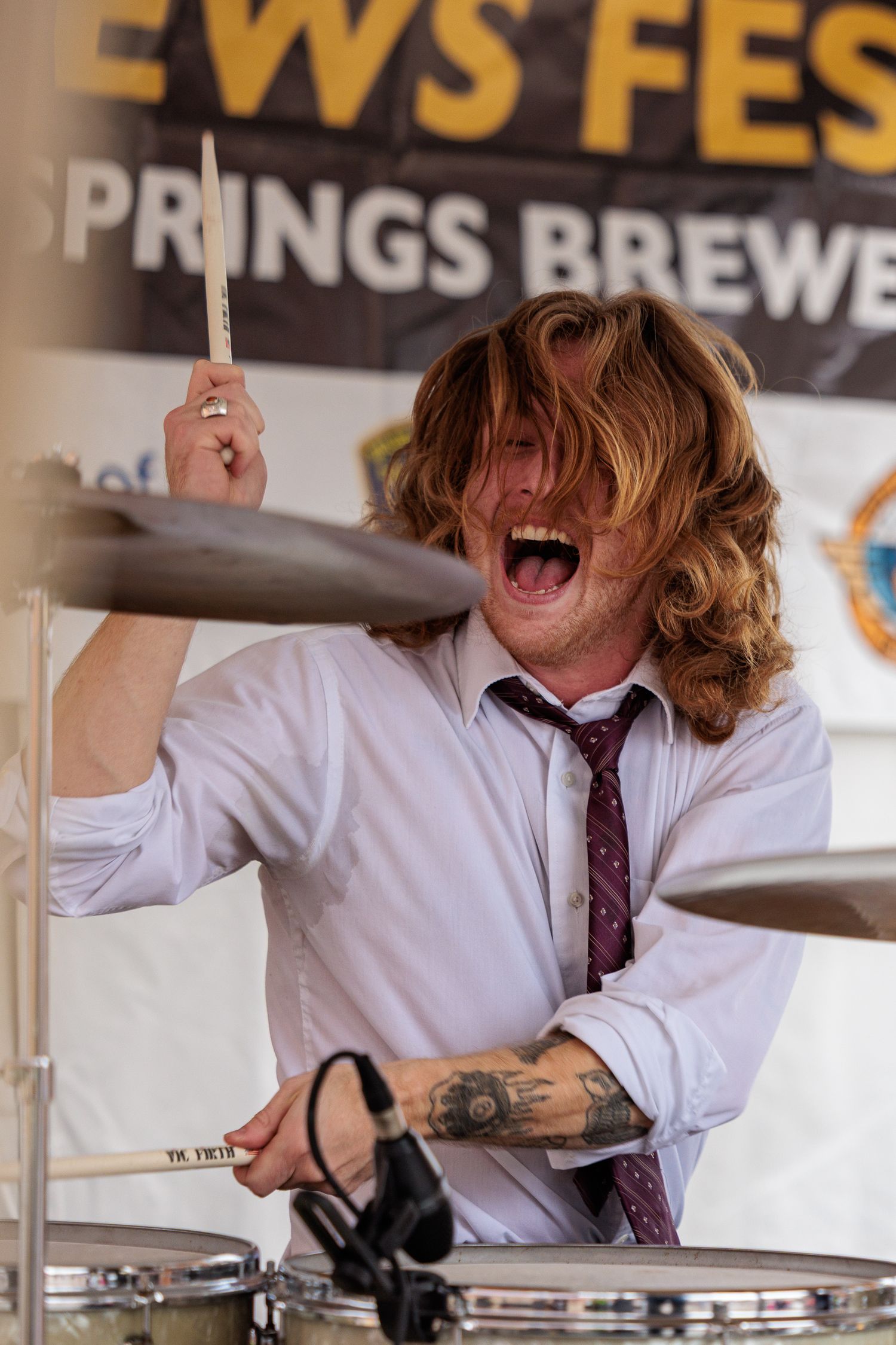 Energetic drummer with long wavy hair plays passionately in a white shirt and tie at an outdoor brewery festival.