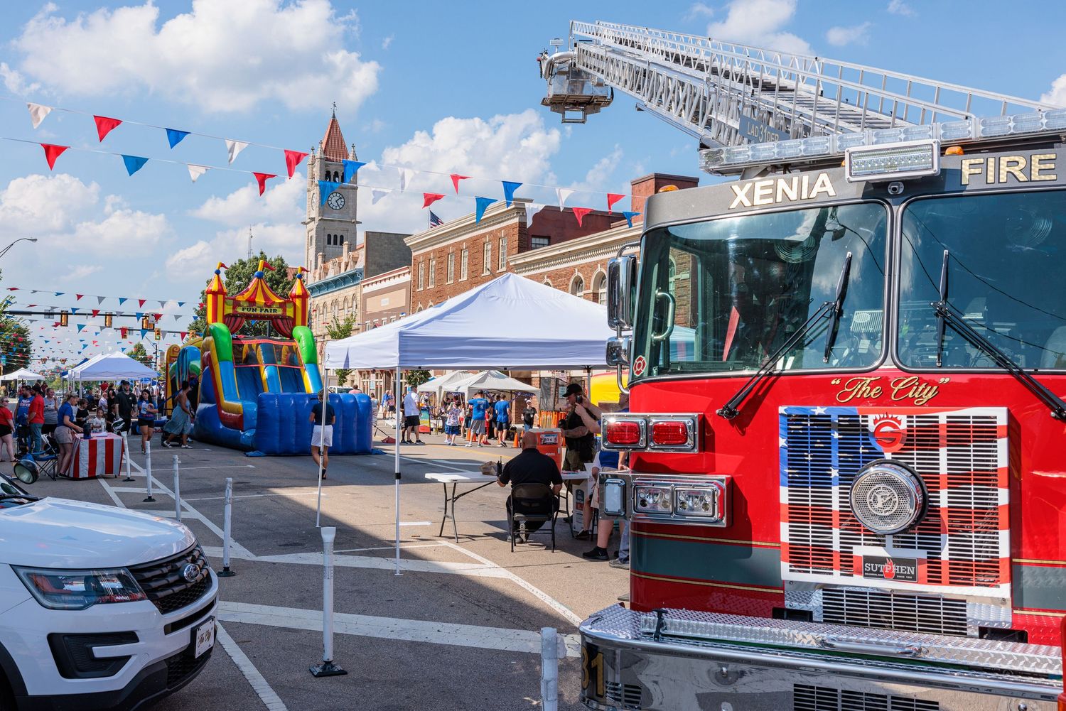 Xenia Fire Department truck at a lively street festival with colorful flags, inflatable bounce house, and vendor tents.