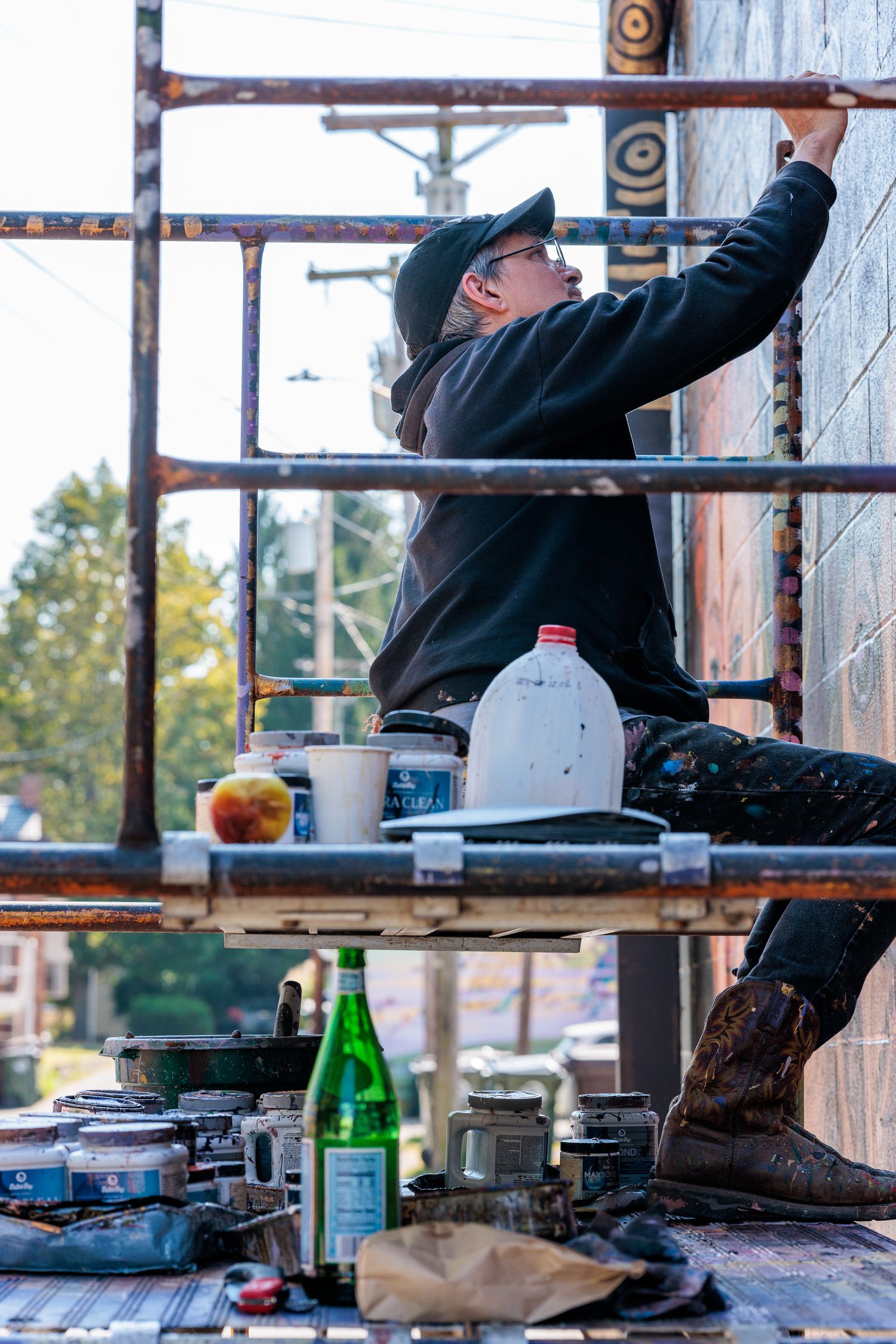 Muralist Pierre Nagley reaching up to paint from scaffolding, paint supplies staged below