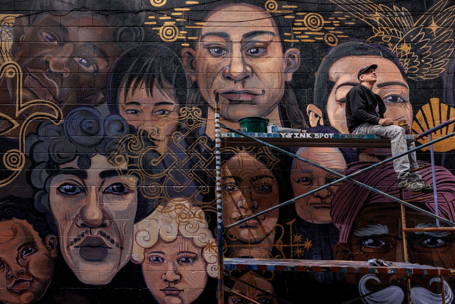 Muralist Pierre Nagley on scaffolding painting a wall mural of many faces for his Immigration series