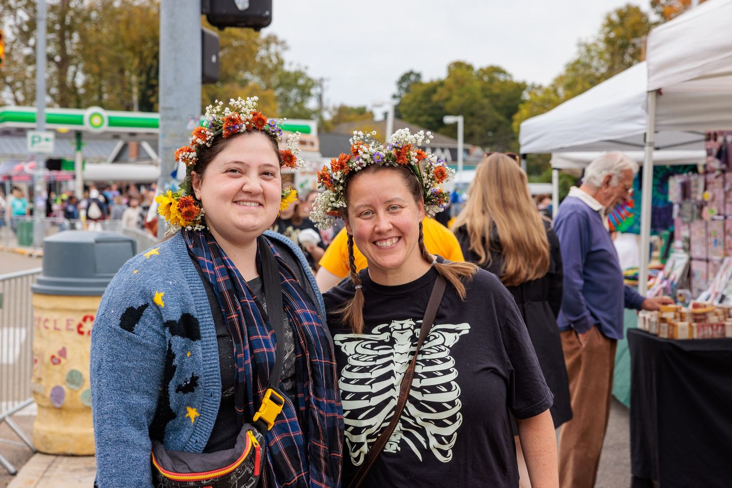 Two smiling women wearing floral crowns pose together at an outdoor market, one in a denim jacket and one in a skeleton rib cage t-shirt.