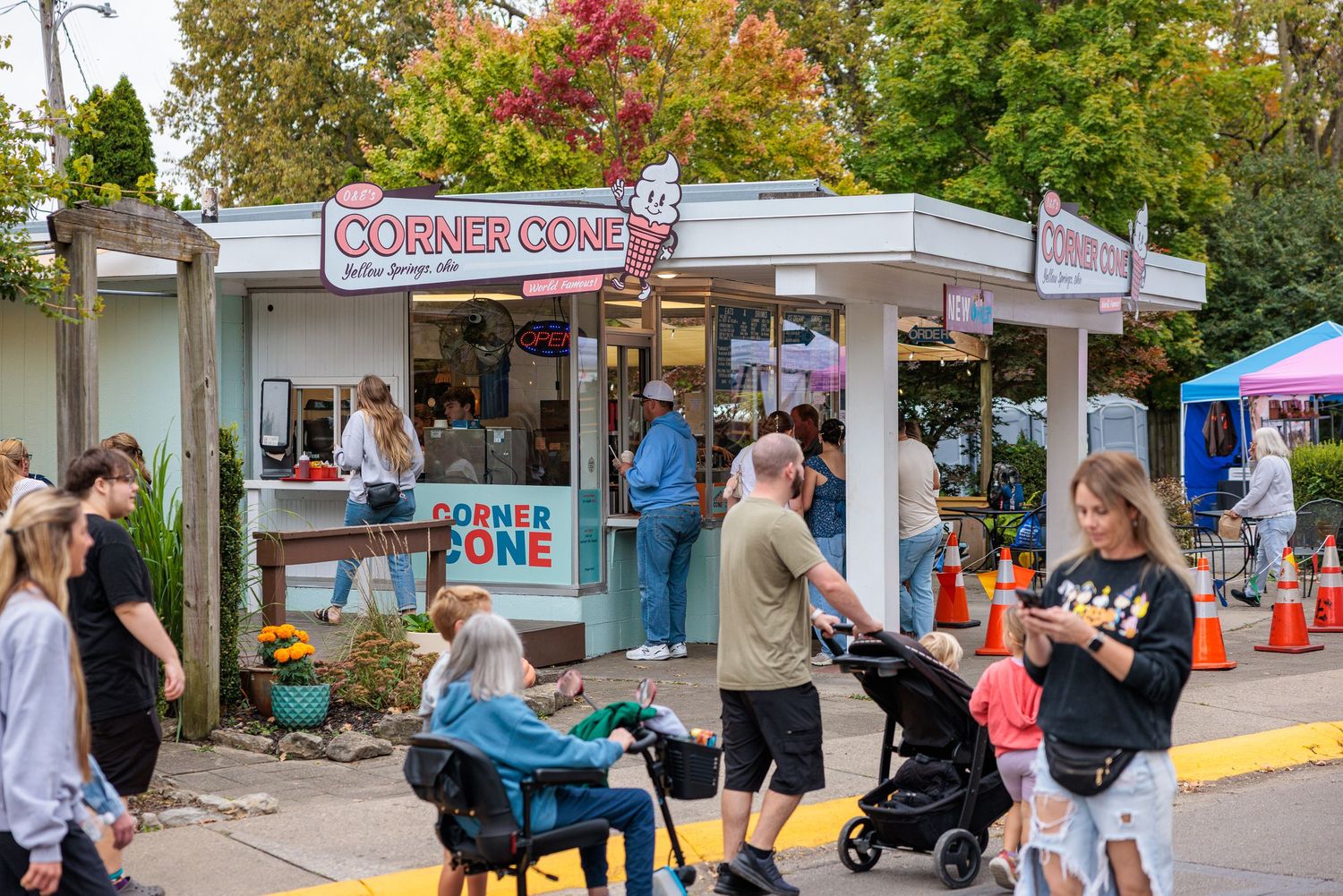 Families and visitors line up outside the Corner Cone ice cream shop on a sunny day, with strollers, orange cones, and autumn trees nearby.
