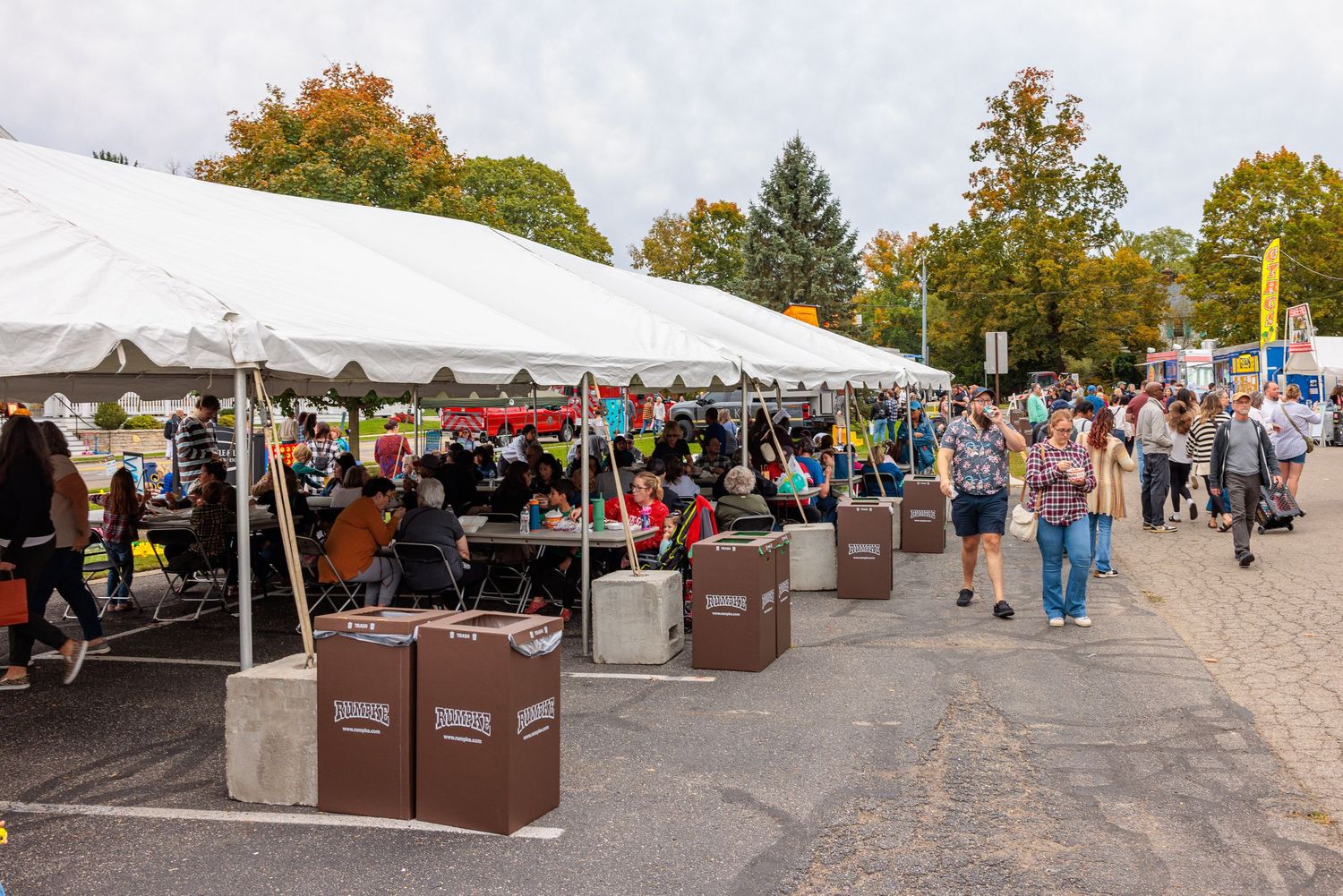 Visitors gather under a large white tent with picnic tables and brown waste bins at an outdoor autumn festival, with colorful trees behind.