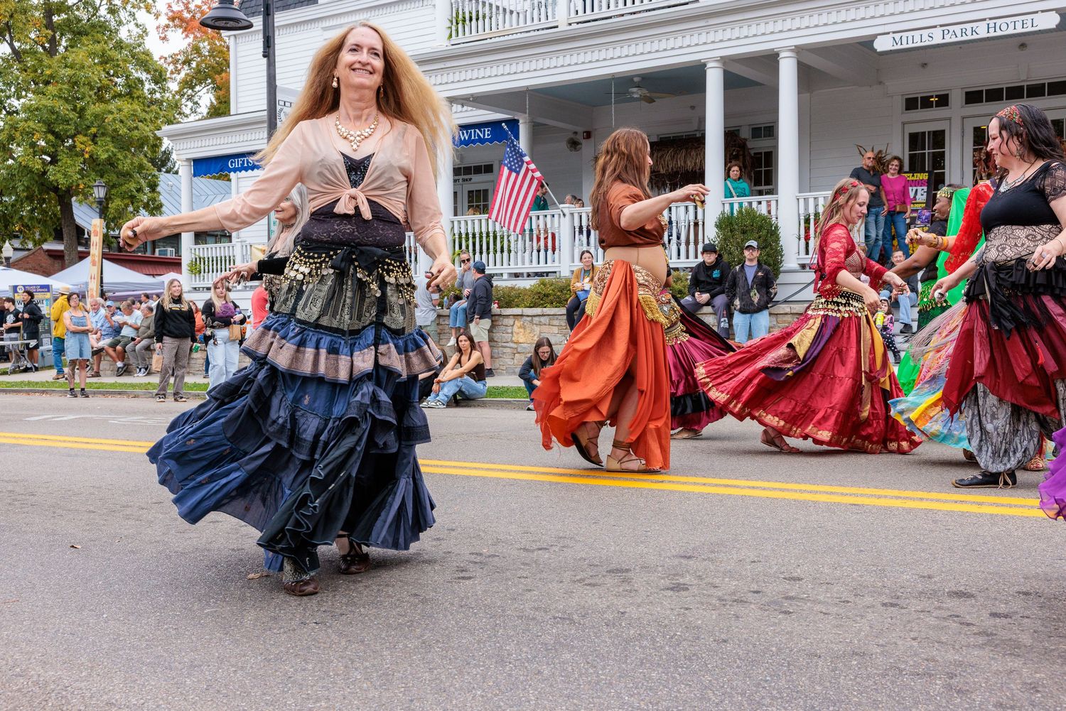 Women in flowing tiered skirts perform a tribal belly dance during a street parade past a white building with an American flag.