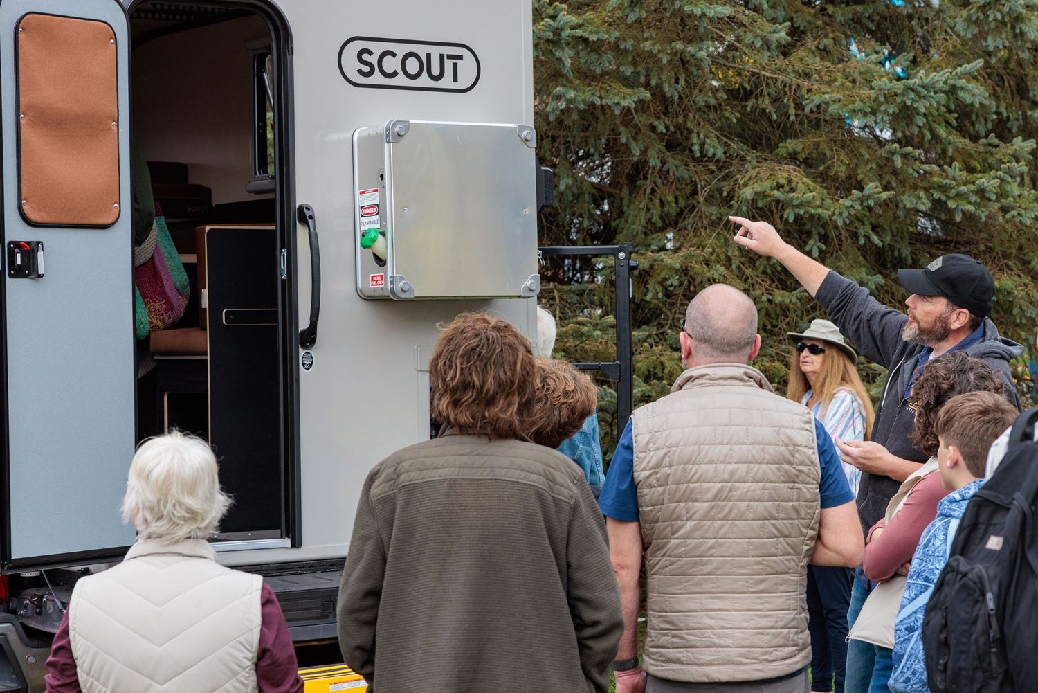 Group of people gathers outside a Scout camper van while a man in a cap points out features during an outdoor tour.