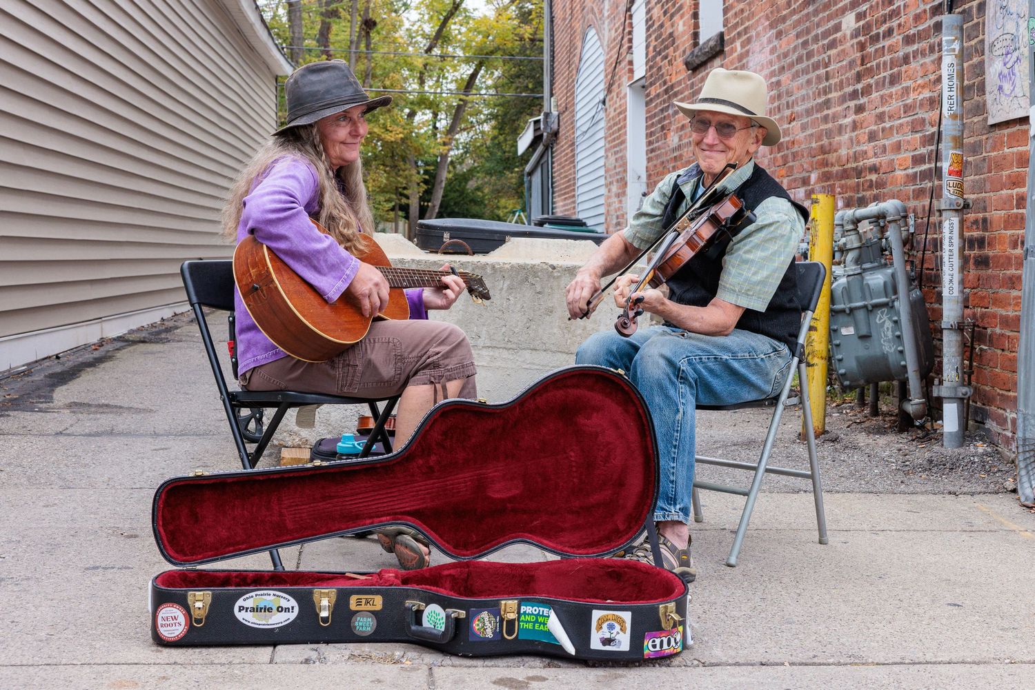 Two street musicians in hats play guitar and fiddle on folding chairs in a brick alley, with an open red instrument case in front.