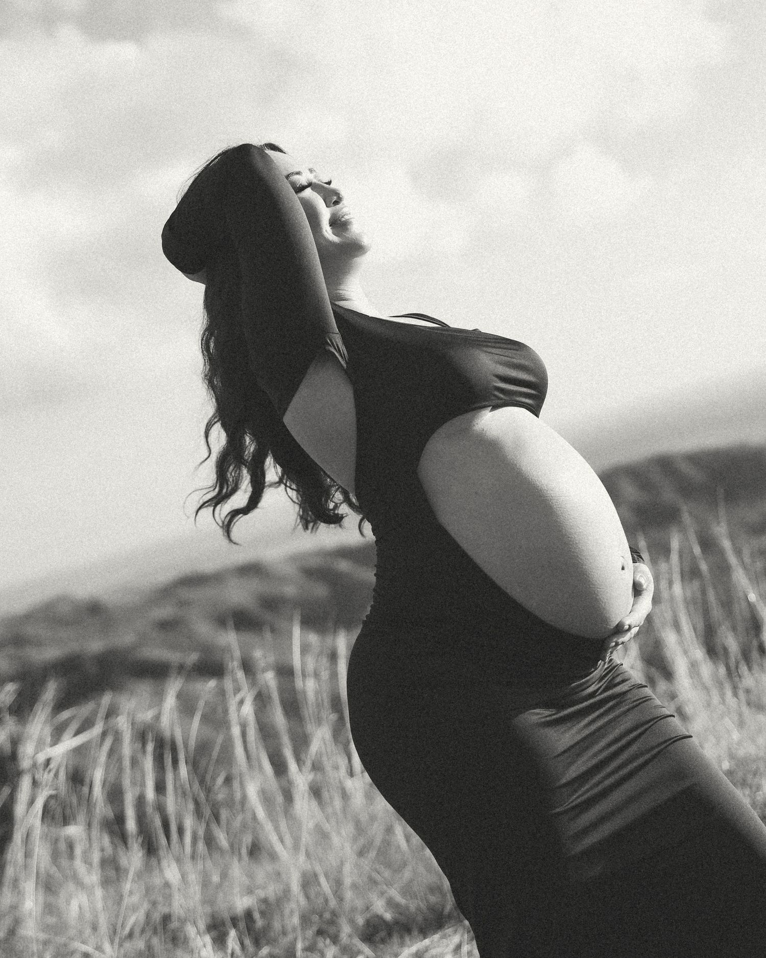 Black and white maternity photo of a pregnant woman in a black dress, posing outdoors in a field with cloudy skies.
