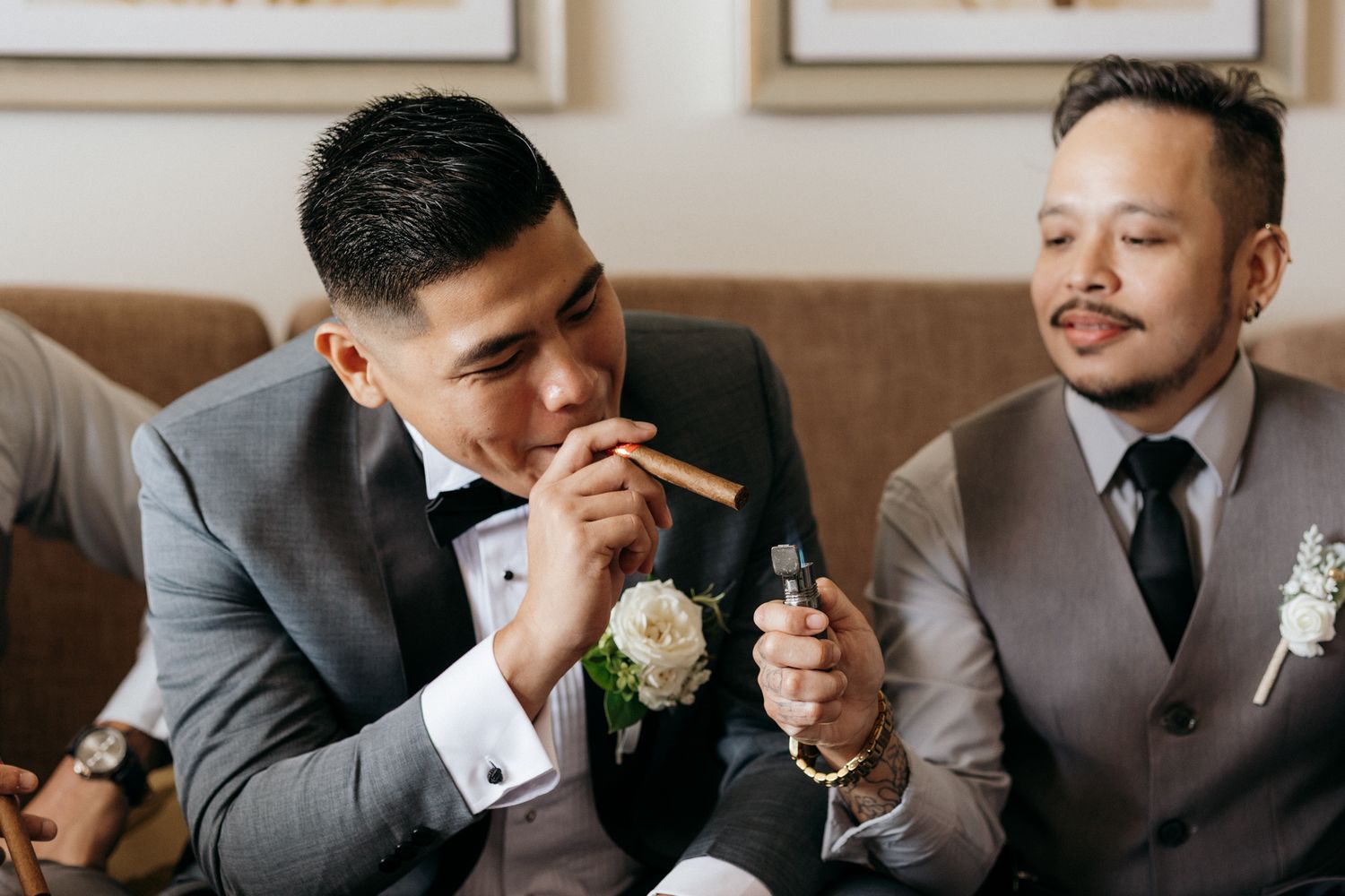 Two groomsmen in suits lighting a cigar together before a wedding ceremony.