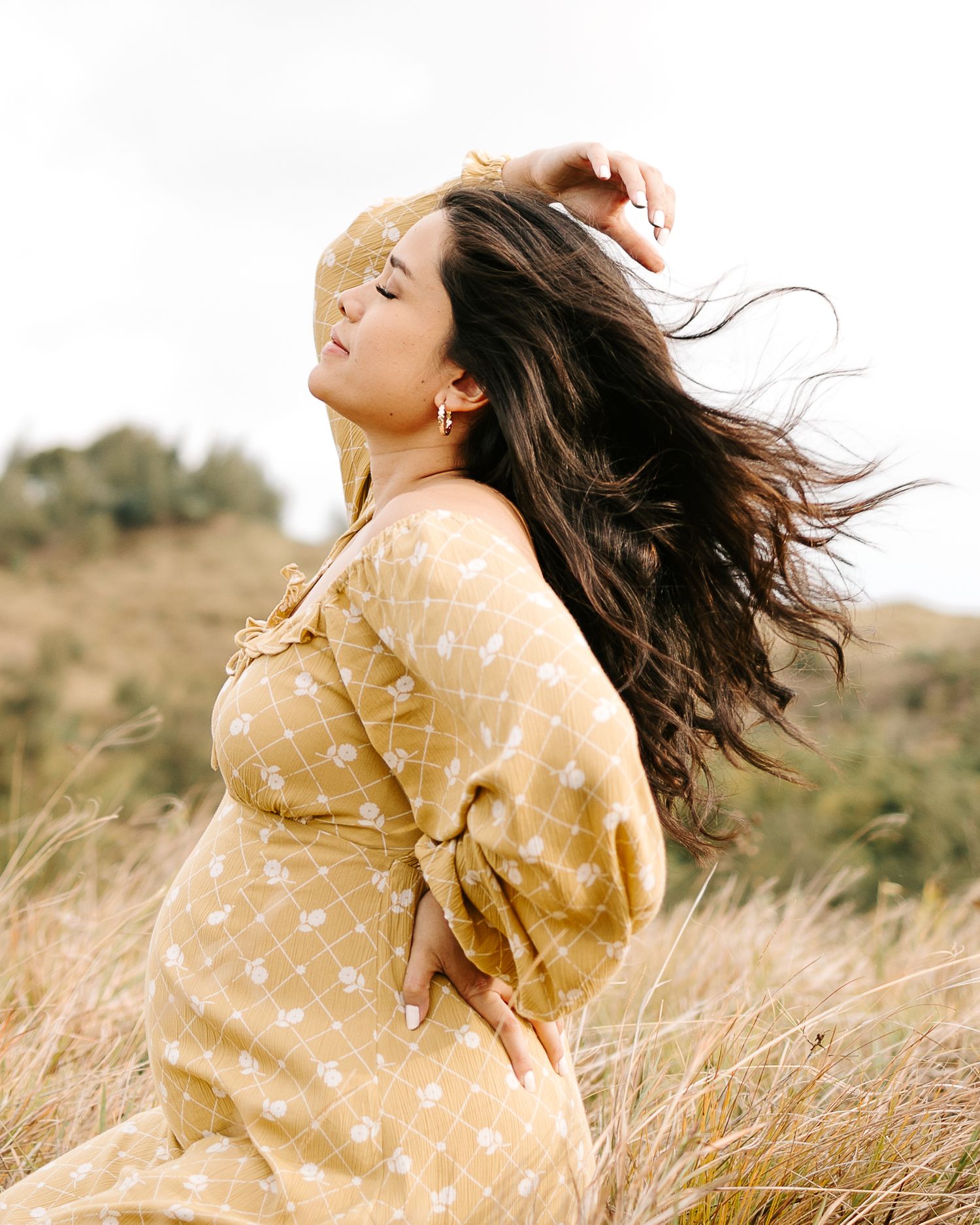 Woman in yellow floral dress poses gracefully in golden grass field, hair flowing in the wind.