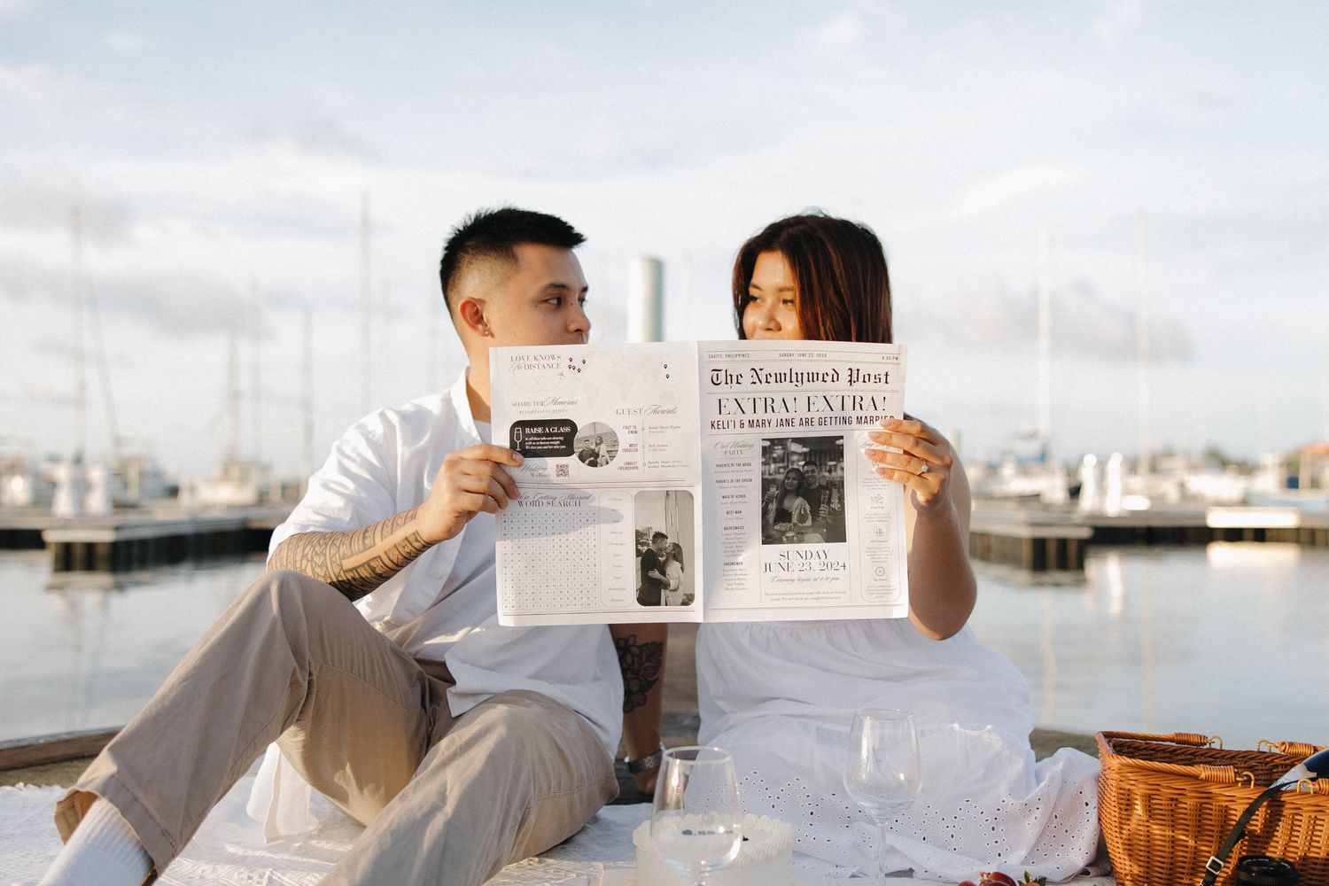 Couple sitting at a marina reading a newspaper together during a romantic outdoor picnic with sailboats in the background.