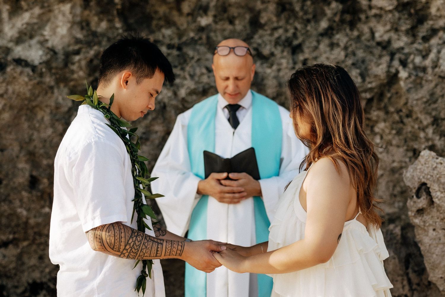 Couple exchanges vows outdoors with officiant in turquoise stole, groom wearing lei and tattoos, bride in white dress.