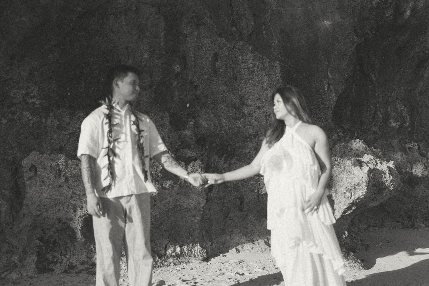 Black and white photo of a couple holding hands on a tropical beach, man wearing a lei, woman in white dress.