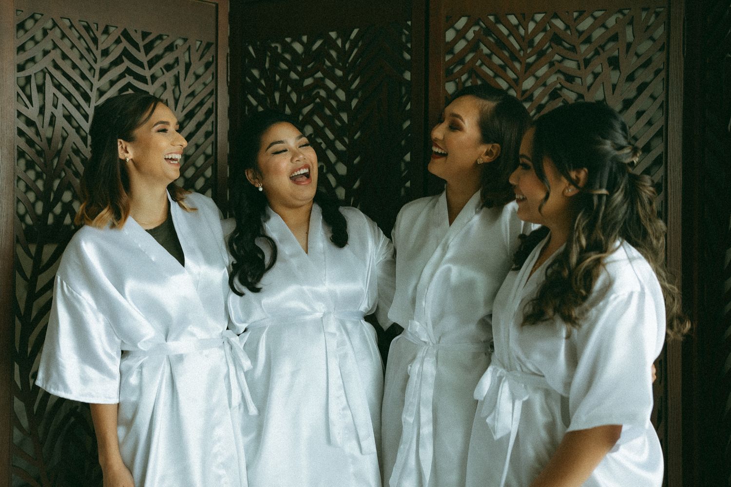 Four women in white satin robes laughing together during bridal party preparations.