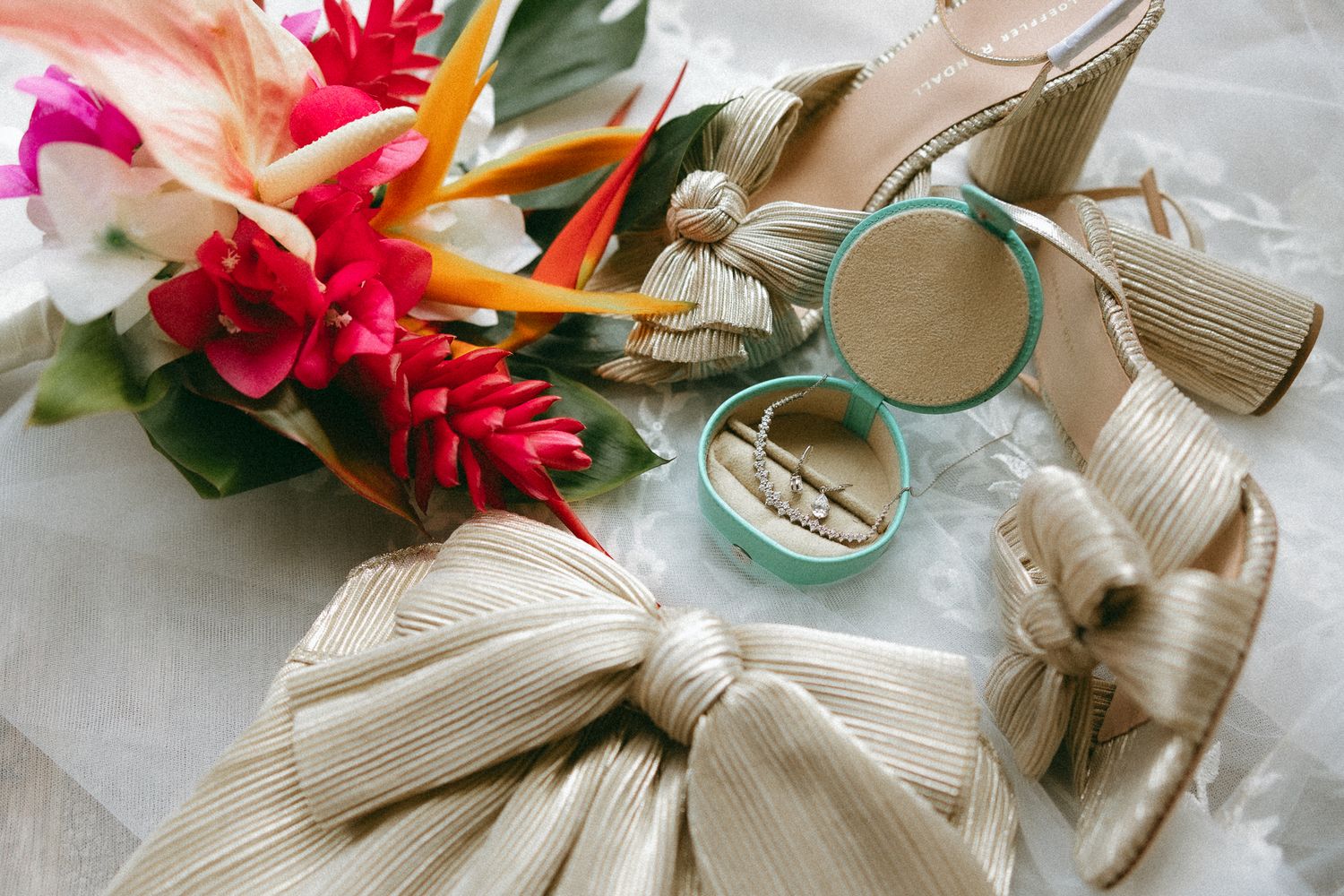 Gold strappy heels with bow detail beside a tropical floral bouquet and turquoise ring box on white surface.