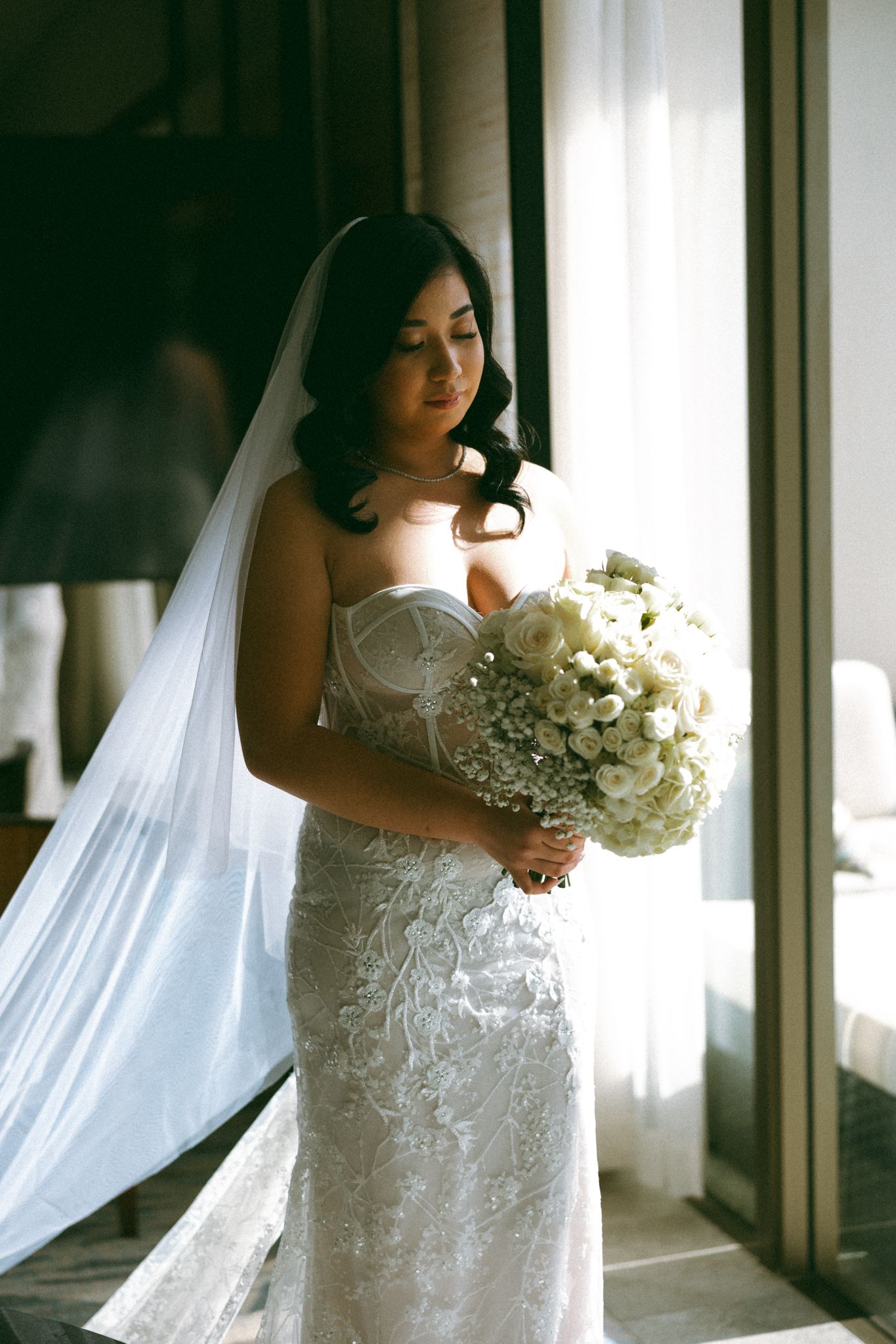 Bride in strapless lace wedding gown and veil holds white floral bouquet near sunlit window.