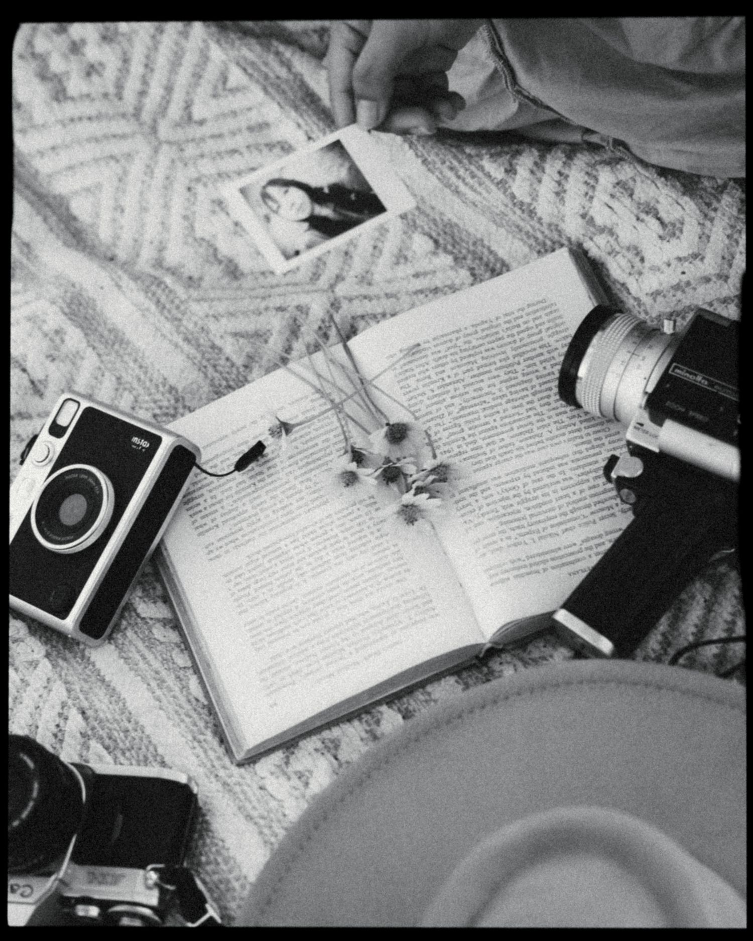 Black and white flat lay of open book, vintage cameras, dried flowers, polaroid photo, and hat on textured blanket.