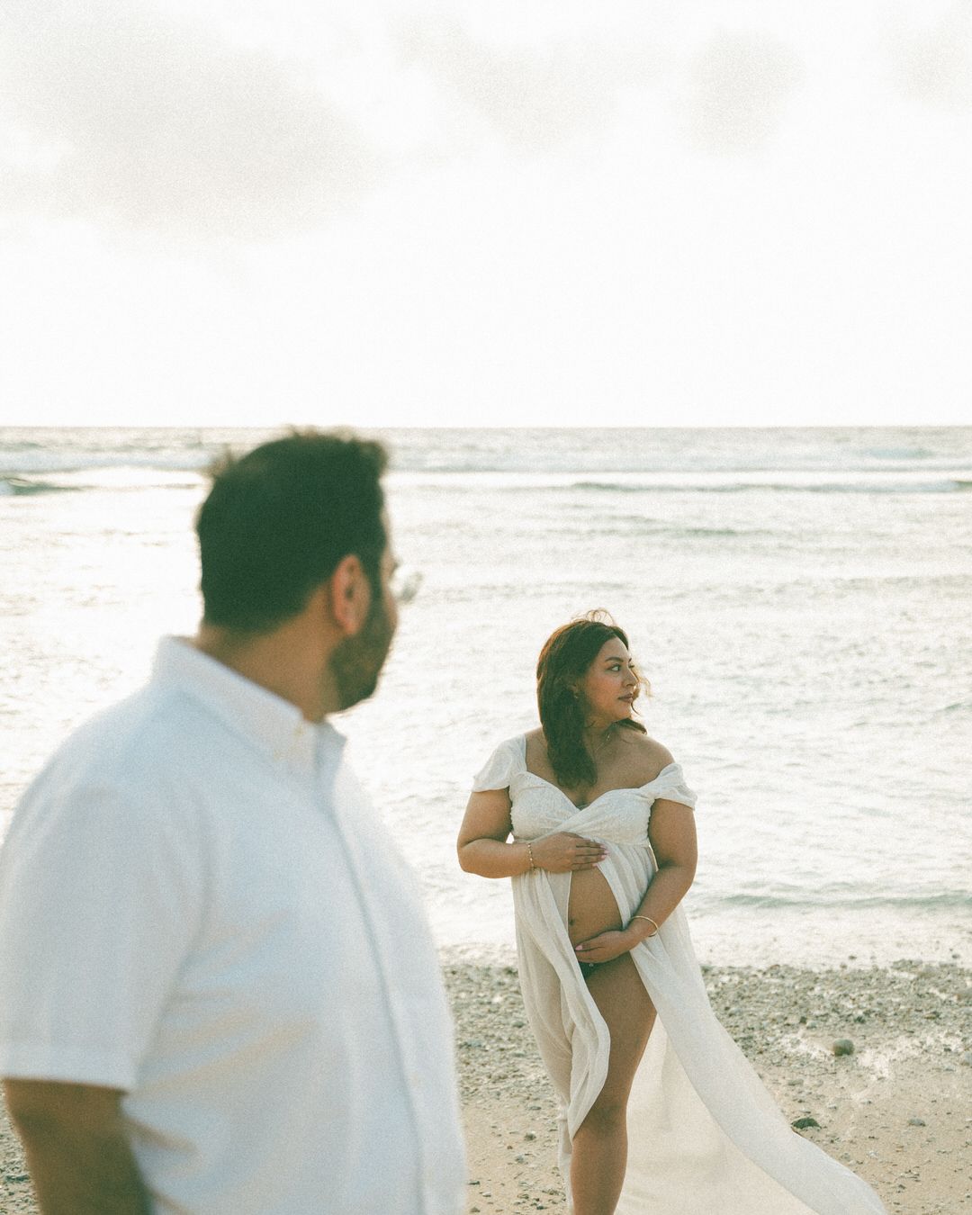 Pregnant woman in white off-shoulder gown poses on beach while man in white shirt stands in foreground.