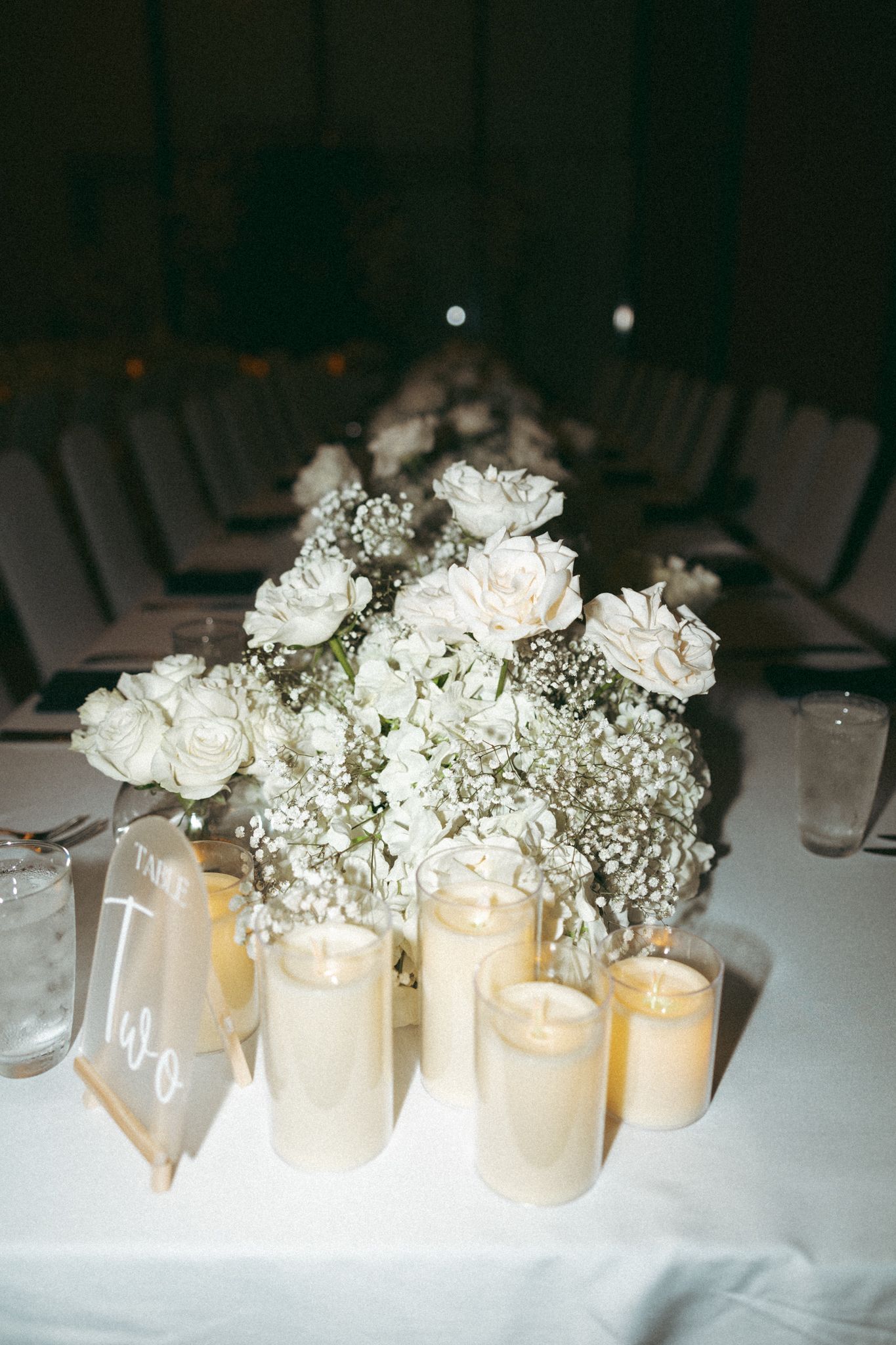 Elegant wedding reception table with white roses, baby's breath centerpiece, and glowing pillar candles on white linen.