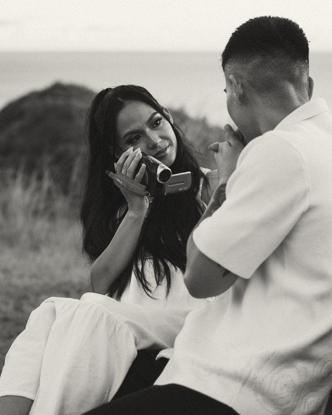 Black and white photo of a couple sitting outdoors, woman holds a camera while man lights her cigarette near the ocean.