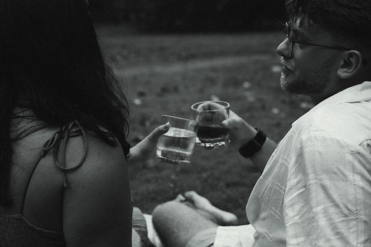 Black and white photo of two people toasting drinks outdoors, man wearing glasses and watch clinking glasses with woman.