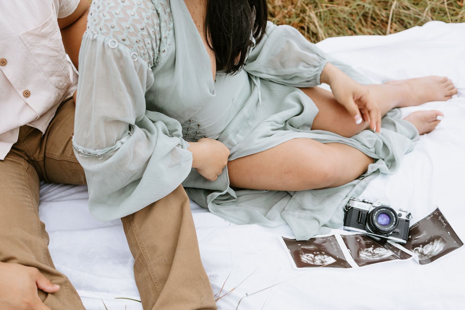 Pregnant couple sitting on white blanket outdoors with ultrasound photos and vintage camera nearby.