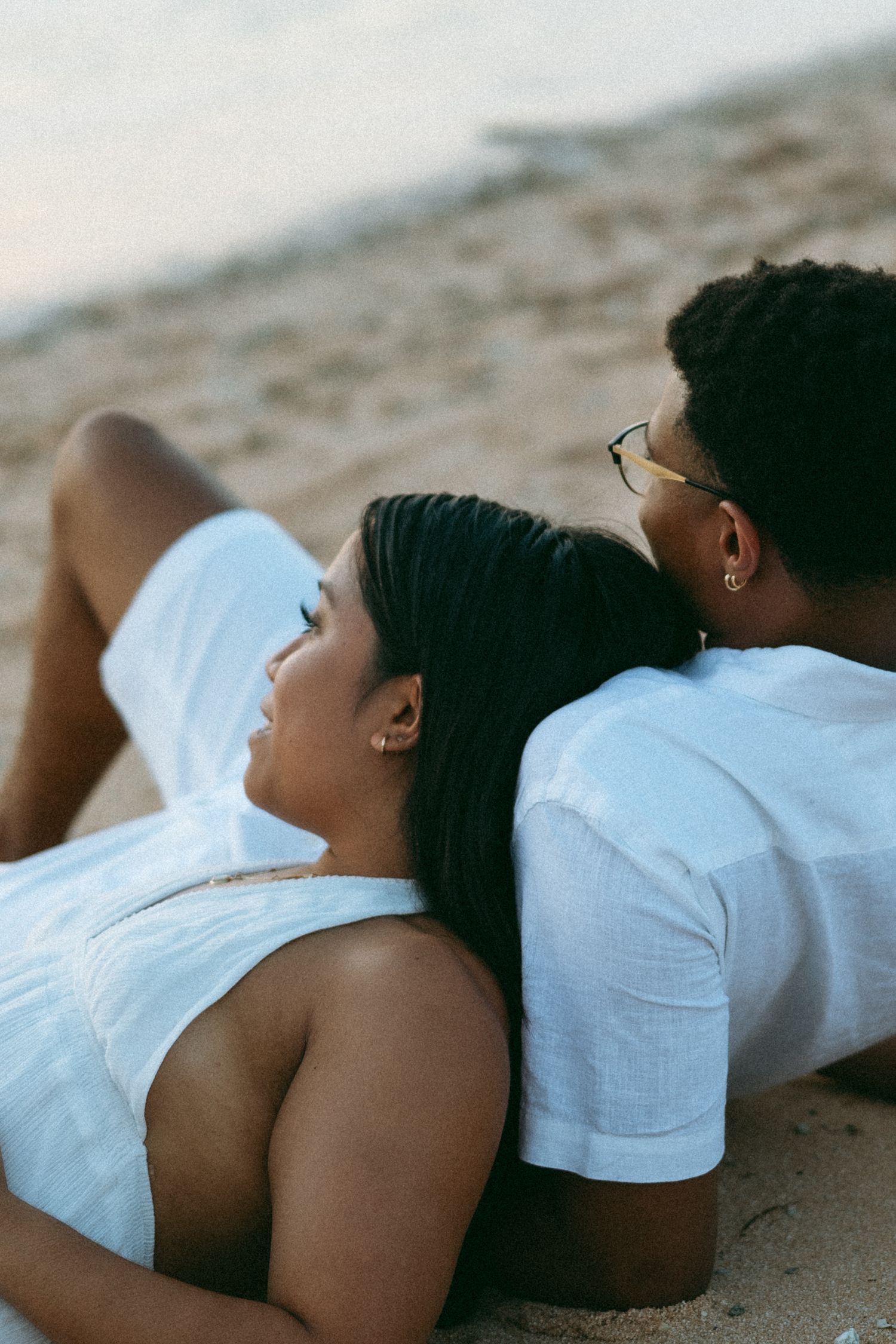 Couple in white outfits relaxing on sandy beach, woman lying back on man's lap gazing up romantically.