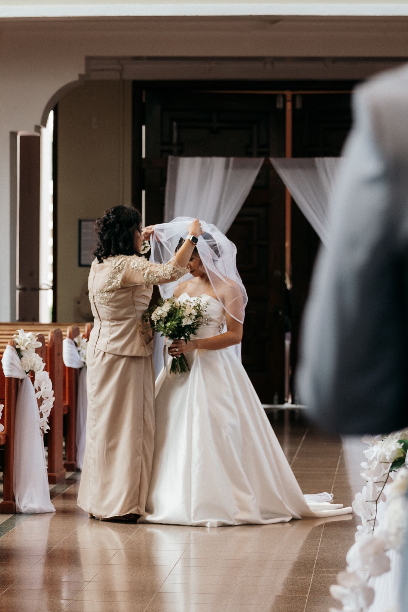 A woman lifts a bride's veil inside a decorated church as the bride holds a bouquet before her wedding ceremony.