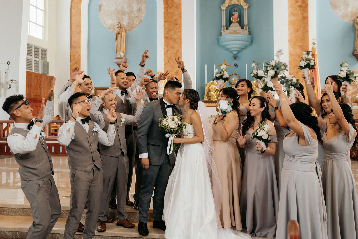 Wedding party celebrating in a blue church, bride and groom surrounded by bridesmaids in gray dresses and groomsmen cheering.