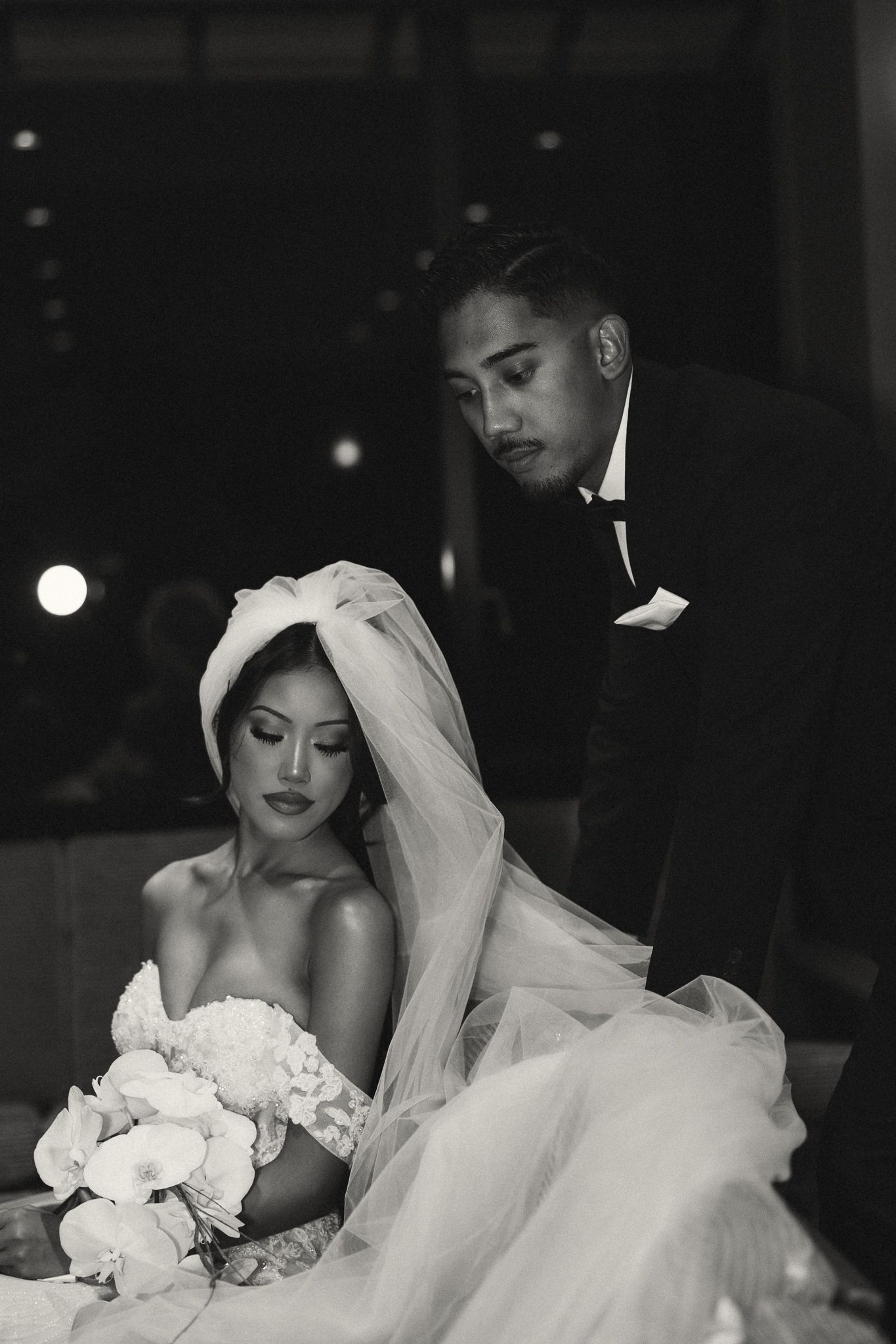 Black and white wedding photo of bride holding white bouquet while groom in tuxedo leans over her tenderly.