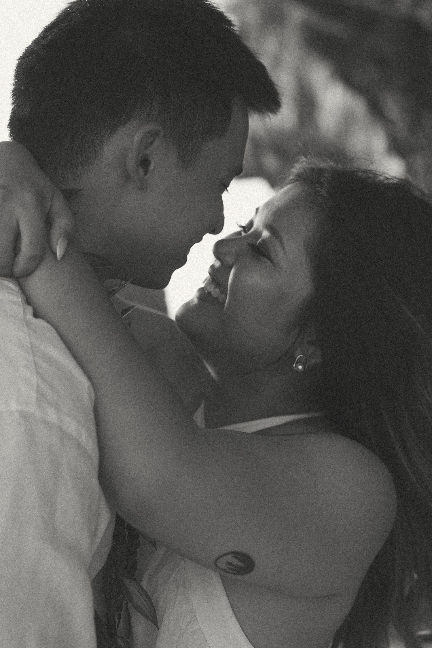 Black and white photo of a smiling couple embracing intimately, gazing at each other in a romantic outdoor setting.