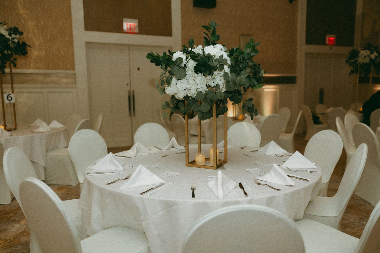 Elegant wedding reception table with white linens, gold centerpiece, greenery, and white floral arrangement in a ballroom.