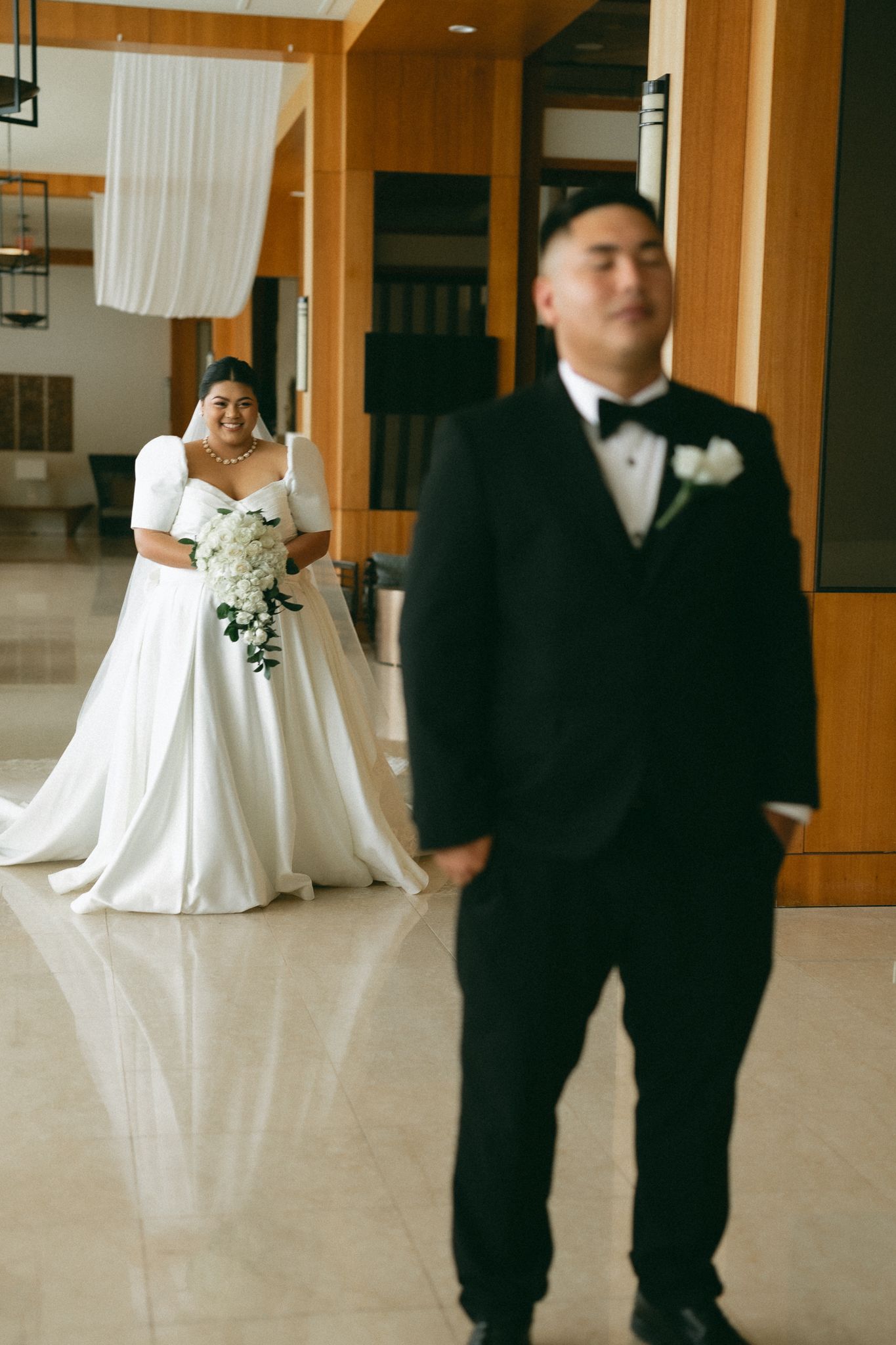 Bride in white gown holds bouquet as groom in black tuxedo stands in foreground during wedding first look.