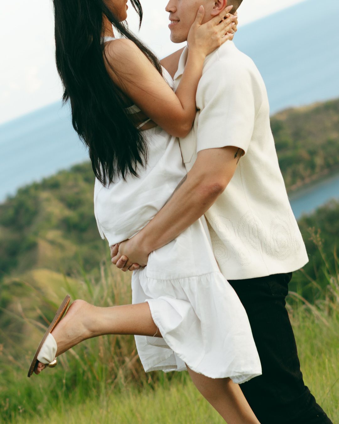 Couple in white outfits embracing romantically on a green hillside with ocean views in the background.