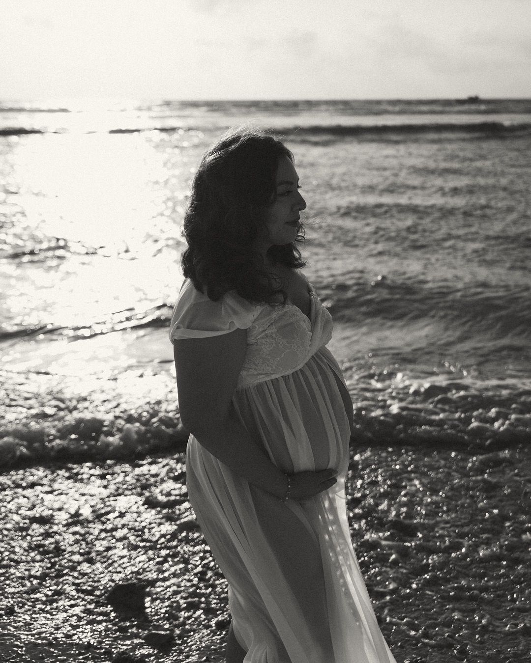 Black and white photo of pregnant woman in white dress standing on beach shore with ocean waves.